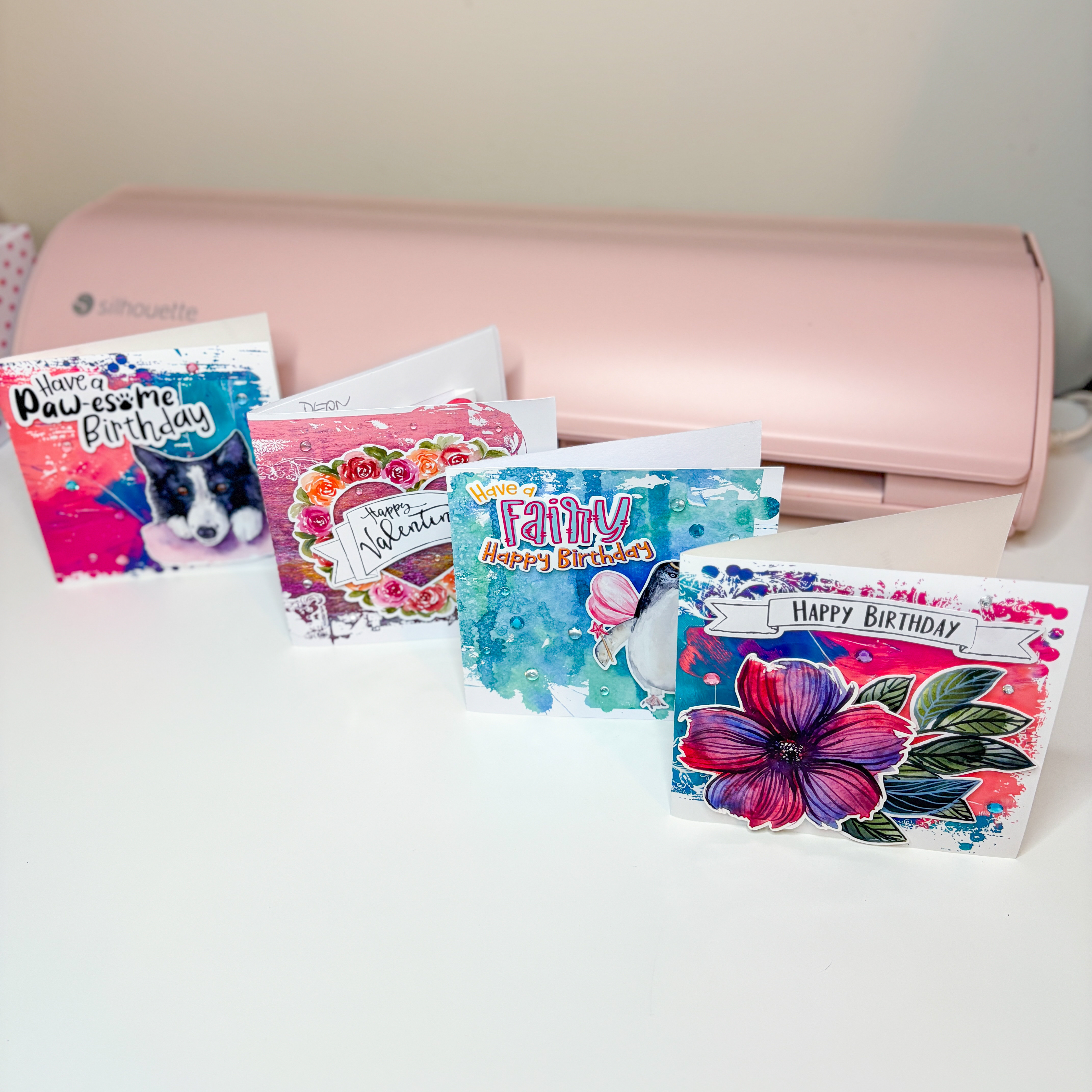 Vibrant handmade Print & Cut birthday and Valentine cards displayed in front of a pink Silhouette cutting machine on a white desk