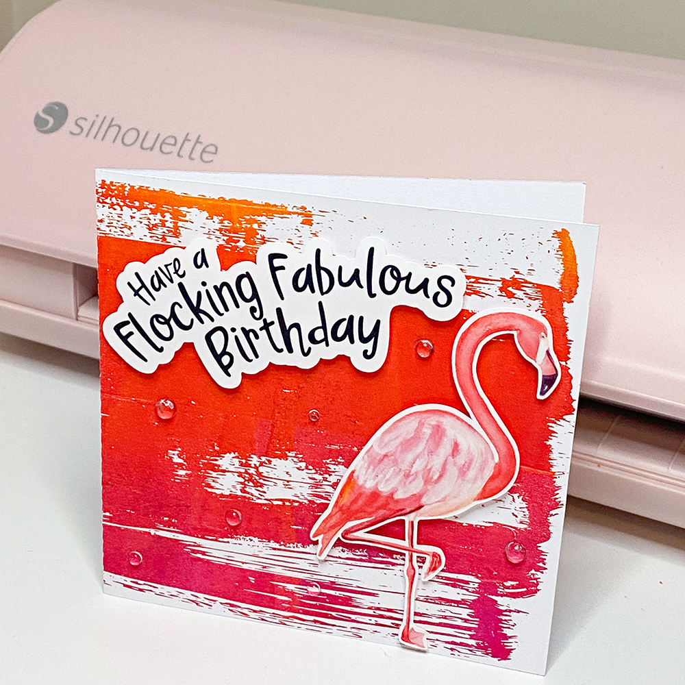 Flamingo-themed handmade birthday card by Clikchic Designs featuring a vibrant red, orange, and magenta monoprint background with grunge edges, overlaid with a pink watercolor-style flamingo and a bold black and white layered sentiment reading "Have a Flocking Fabulous Birthday," displayed in front of a pink Silhouette cutting machine.